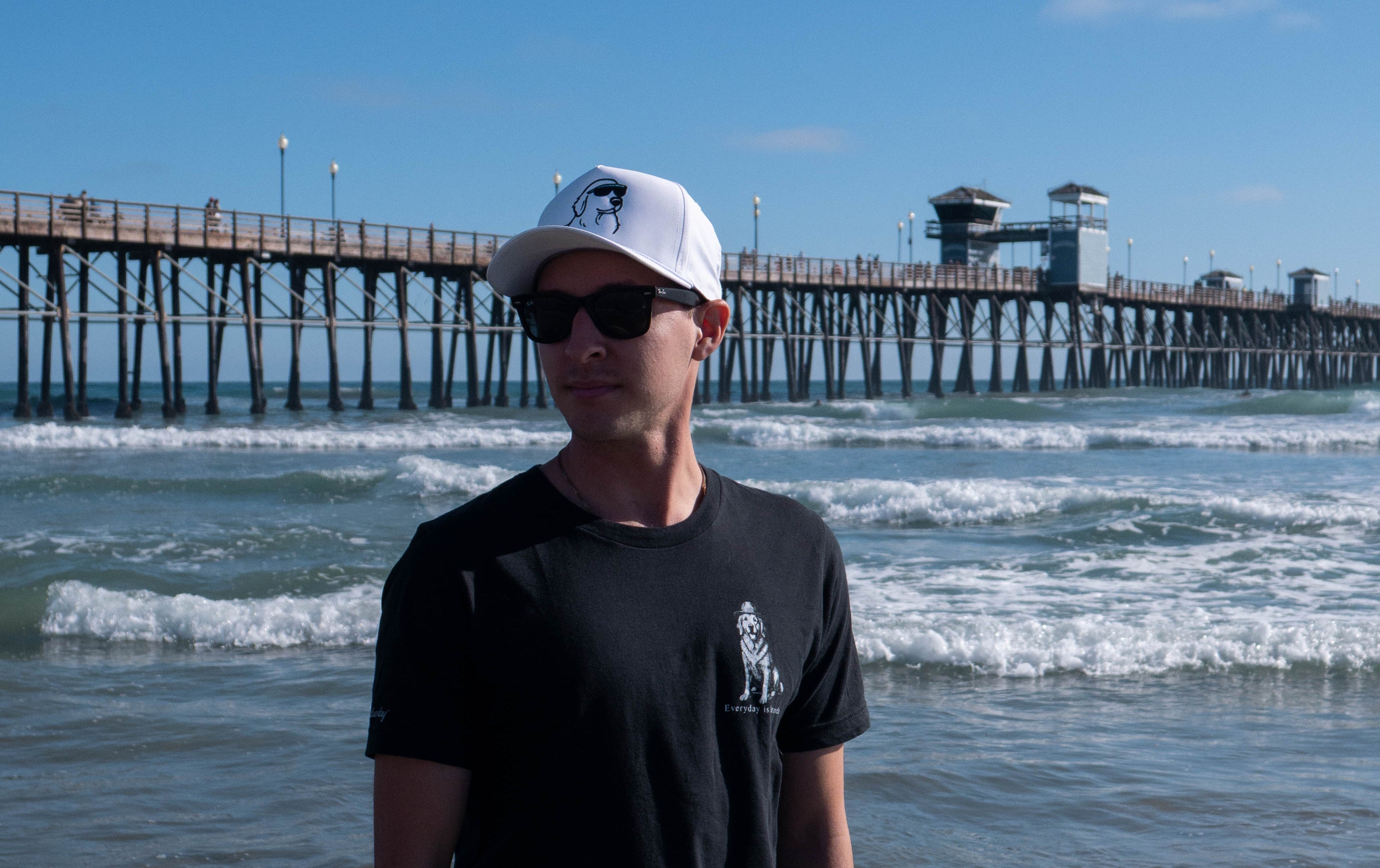 A man standing on a beach in front of the ocean and a pier, wearing a black t-shirt with a small white golden retriever graphic, a white baseball cap with a dog wearing sunglasses, sunglasses, and light green shorts.