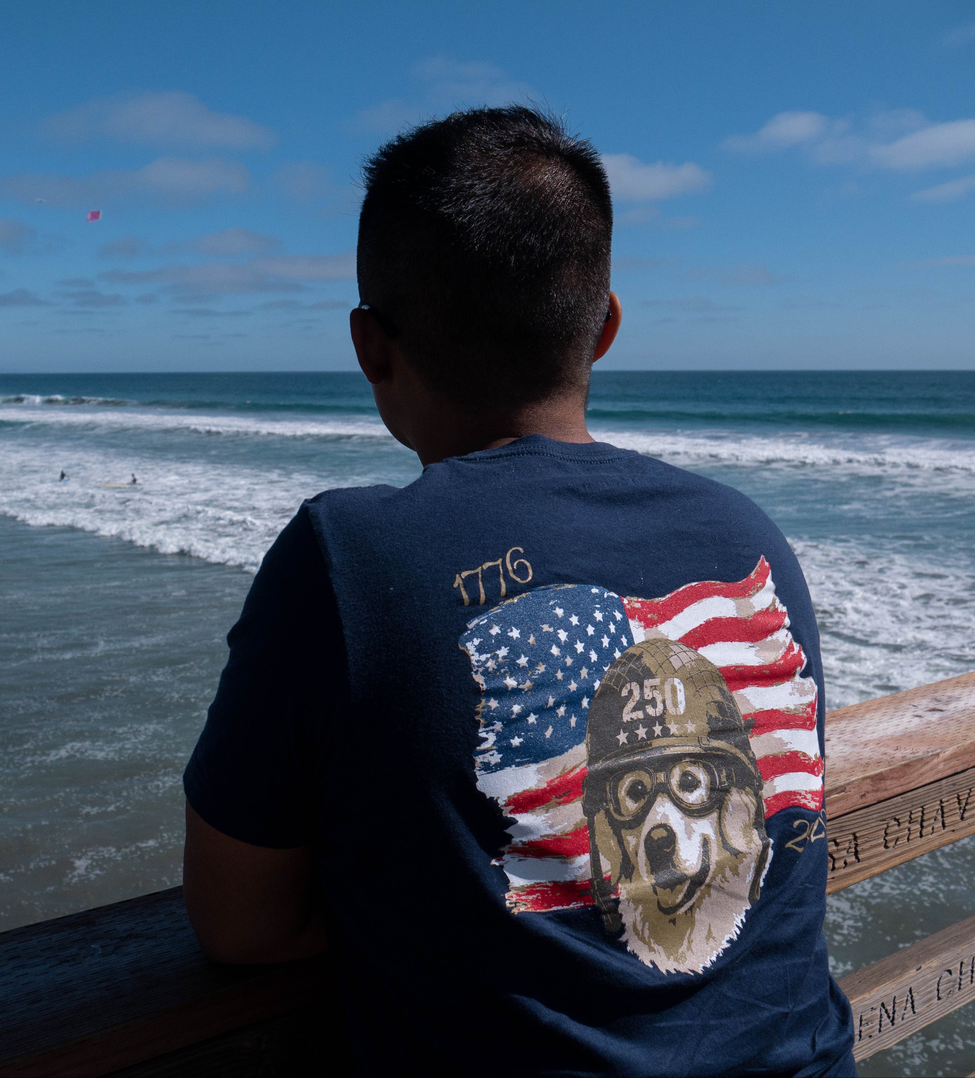 A man seen from behind sitting on a wooden boardwalk railing, looking out at the ocean waves under a blue sky. He is wearing a navy blue t-shirt with a large graphic on the back featuring a dog in a military helmet and an American flag.